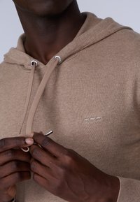 Hoodie en polaire beige avec une finition texturée, présentant des œillets en métal argenté, un cordon de serrage et un subtil détail brodé sur la poitrine.