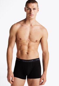 Svarte boxershorts med logo på midjen, laget av glatt stoff. Passformen er tett, med sømdetaljer. Modellen står foran en hvit bakgrunn.