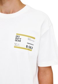 Hvid bomulds t-shirt med et rektangulært grafisk design med sort og guld tekst, der står "OFF DUTY GEAR" samt numeriske detaljer. Kortærmet.