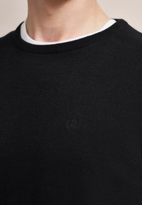 Nahaufnahme einer Person, die einen schwarzen Rundhals-Pullover über einem weißen Unterhemd mit einem dezent gestickten Logo auf der Brust trägt.