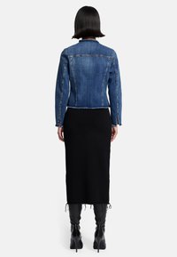 Dunkelblaue Jeansjacke mit cropped Schnitt und fransigen Kanten; getragen über einem schwarzen knielangen Kleid und schwarzen kniehohen Stiefeln.