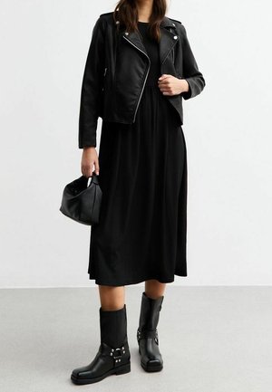 Kobieta ubrana w czarną skórzaną kurtkę, czarną sukienkę midi, czarne kozaki do połowy łydki z srebrnymi klamrami, trzymająca czarną torebkę.
