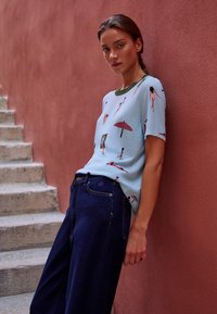 Kobieta z warkoczami opiera się o fakturowaną różową ścianę, ubrana w jasnoniebieską koszulę z nadrukiem pływaka i parasola oraz ciemnoniebieskie szerokie dżinsy.