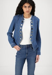 Jaqueta de denim azul com um design ajustado, apresentando botões metálicos à frente, combinada com uma blusa branca e jeans de cintura alta.