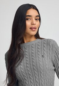 Pullover grigio a maglia con collo rotondo. Presenta un motivo testurizzato e un piccolo logo sul petto. Tessuto morbido con una forma aderente.