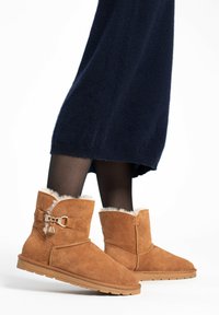 Botas de tobillo de ante marrón con forro blanco esponjoso, con un acento de hebilla en tono dorado, suela plana y diseño de punta redonda.