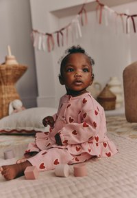 Rosa klänning med röda hjärtmönster, med volangkant och knappar framtill. Barn sitter på en texturerad matta, omgiven av färgglada klossar.