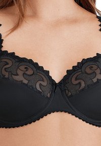 Reggiseno in pizzo nero con una parte superiore trasparente decorata con un motivo a spirale, bordi scallop e una coppa opaca liscia. Include un dettaglio a fiocco centrale.