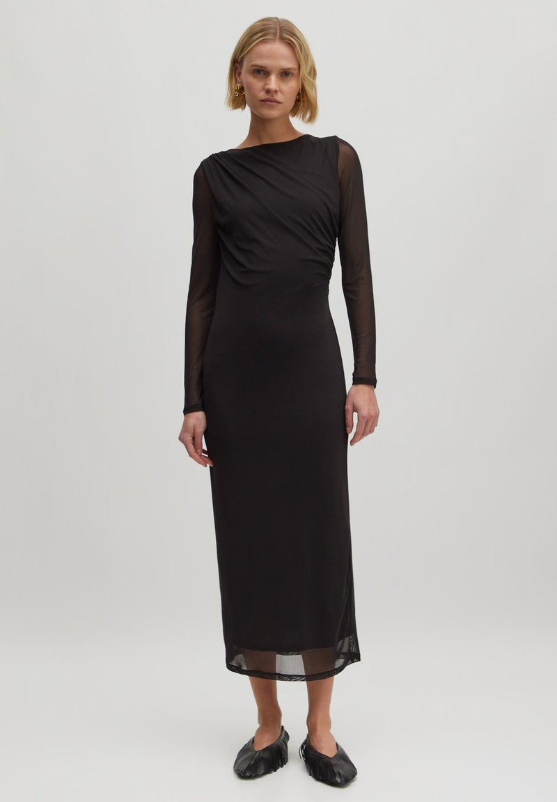 Schwarzes Langarmkleid mit tailliertem Schnitt, das mit Drapierungen, transparenten Ärmeln und einem fließenden Saum versehen ist. Akzentuiert mit subtilen Texturen.