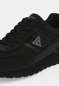Gros plan sur une basket noire Guess avec un laçage et le logo triangulaire Guess sur la partie latérale.