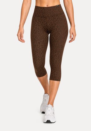 Femme portant des leggings courts imprimé léopard marron et des chaussures de sport blanches marchant sur un fond uni.