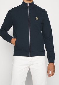 Marinblå zip-tröja med hög krage, ribbade ärmslut, två sidofickor och en liten logotyp-patch på vänster bröst. Mjuk textur.