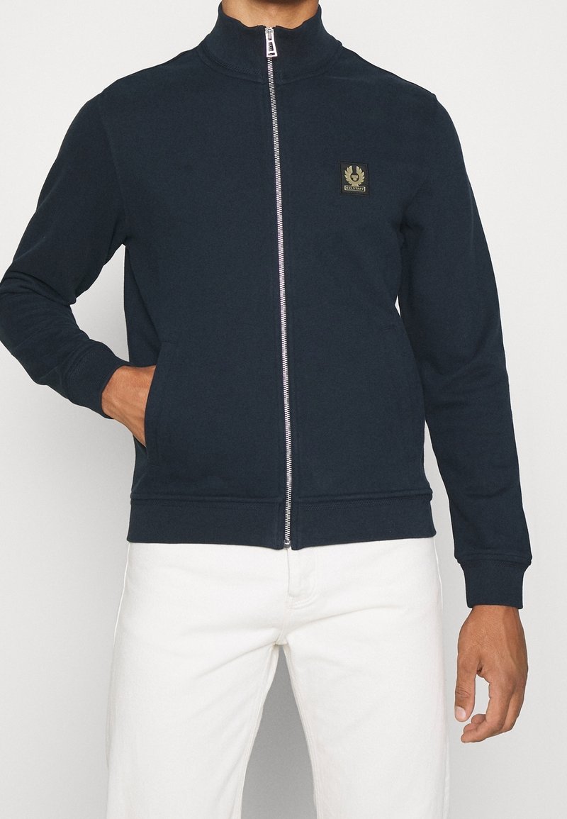 Marinblå zip-tröja med hög krage, ribbade ärmslut, två sidofickor och en liten logotyp-patch på vänster bröst. Mjuk textur.