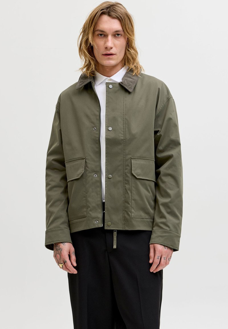 Mladý muž s blond vlasmi po plecia, oblečený v olivovo zelenej bunde s náprsními vreckami, bielom tričku a čiernych nohaviciach, stojaci pred jednoduchým pozadím.