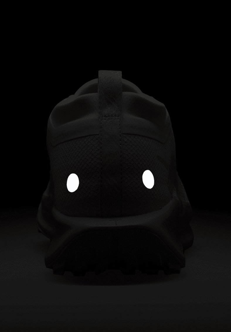 Zapatilla deportiva negra con un upper de malla texturizada, que presenta acentos reflectantes blancos y una suela resistente, vista desde la parte trasera sobre un fondo oscuro.