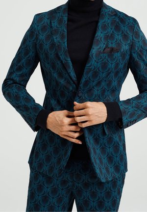 Mann knöpft eine teal gemusterte Anzugjacke über einem schwarzen Rollkragenpullover, mit passenden gemusterten Hosen und schwarzem Einstecktuch.