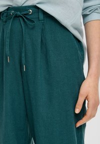 Turkusowe lniane spodnie z elastycznym pasem, z fakturowanym wykończeniem, plisami i sznurkami zakończonymi srebrem; ręka częściowo oparta na pasie.