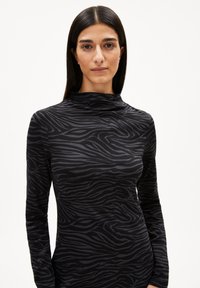 Femme aux longs cheveux bruns portant un haut à manches longues noir et gris foncé à rayures zèbres avec un col montant, faisant face à l'avant sur un fond blanc.