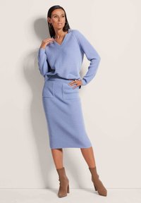 Hellblaue gerippte Strickpullover- und Midikleider-Kombination, mit V-Ausschnitt, langen Ärmeln und zwei aufgenähten Taschen am Rock. Ergänzt durch braune Sockenschuhe.