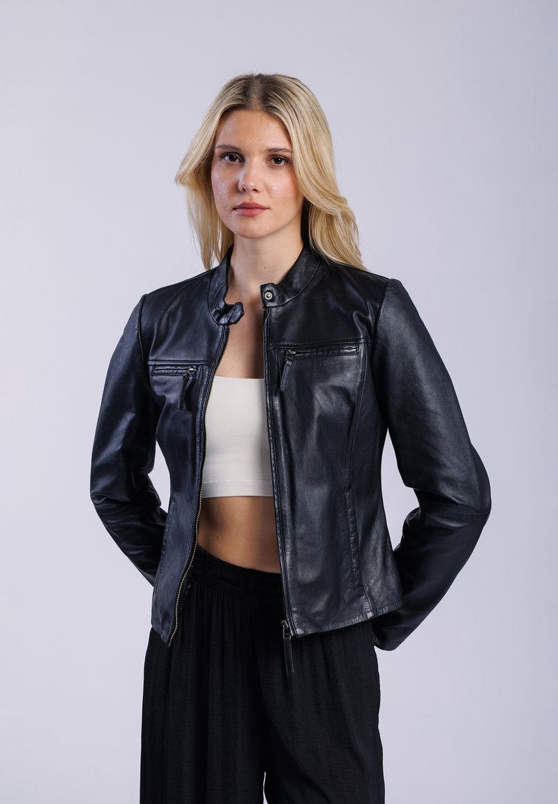 Zwart leren jack met een kraag en een rits aan de voorkant. Heeft twee ritszakken op de borst en een gladde textuur. Draagt over een witte crop top.