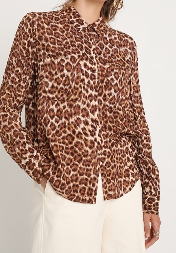 Femme portant une chemise marron à manches longues avec des boutons, imprimé léopard et col, et un pantalon blanc, main dans la poche.