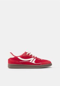 Baskets rouges basses avec des rayures blanches sur les côtés, un patch blanc au talon, des lacets blancs et une semelle marron sur un fond clair uni.