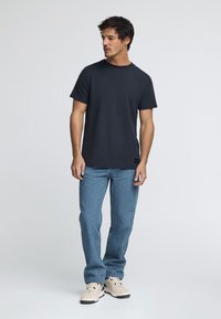 Camiseta negra de cuello redondo, de mangas cortas, combinada con jeans de color azul claro. La camiseta tiene un pequeño parche de logo en el dobladillo, con textura de tela de algodón.