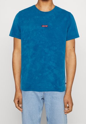 Mand iført en blå batikmønstret Levi's t-shirt og lyseblå jeans, stående mod en ensfarvet hvid baggrund.