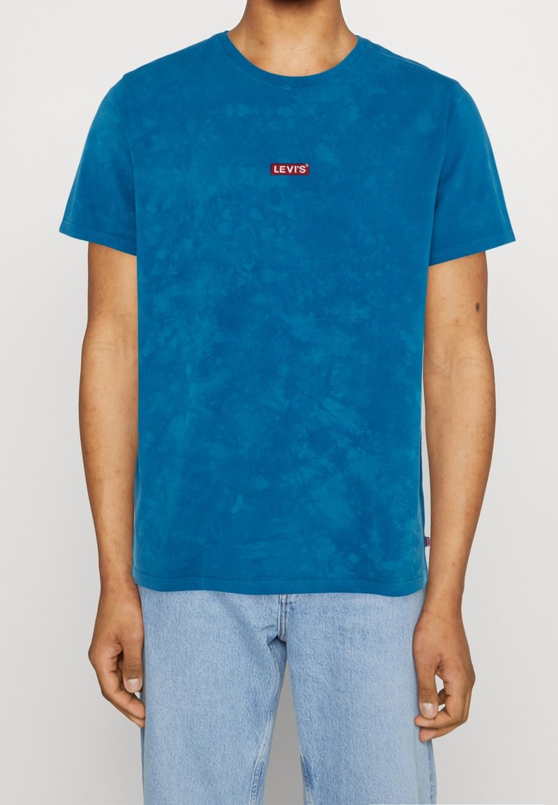 Homme portant un t-shirt Levi's tie-dye bleu et un jean bleu clair, debout devant un fond blanc uni.