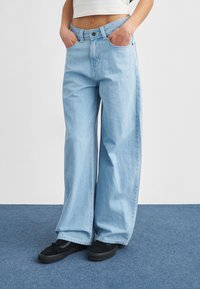 Άτομο που φοράει φαρδιά ανοιχτό μπλε τζιν και μαύρα αθλητικά παπούτσια, στέκεται σε μπλε χαλί με τα χέρια στις τσέπες, κομμένο στη μέση.