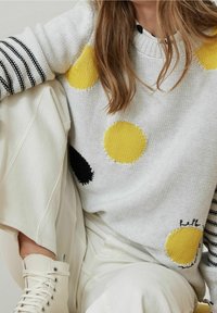 Maglione lavorato a maglia di colore bianco con grandi motivi circolari gialli e neri, dotato di maniche a righe bianche e nere e cuciture decorative.