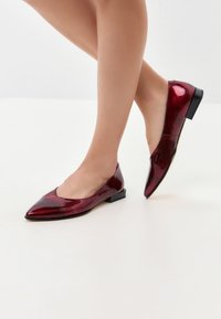 Ballerines en cuir verni rouge avec un bout pointu et un petit talon carré. Texture lisse avec une finition brillante. Vue de côté montrant le placement du pied.