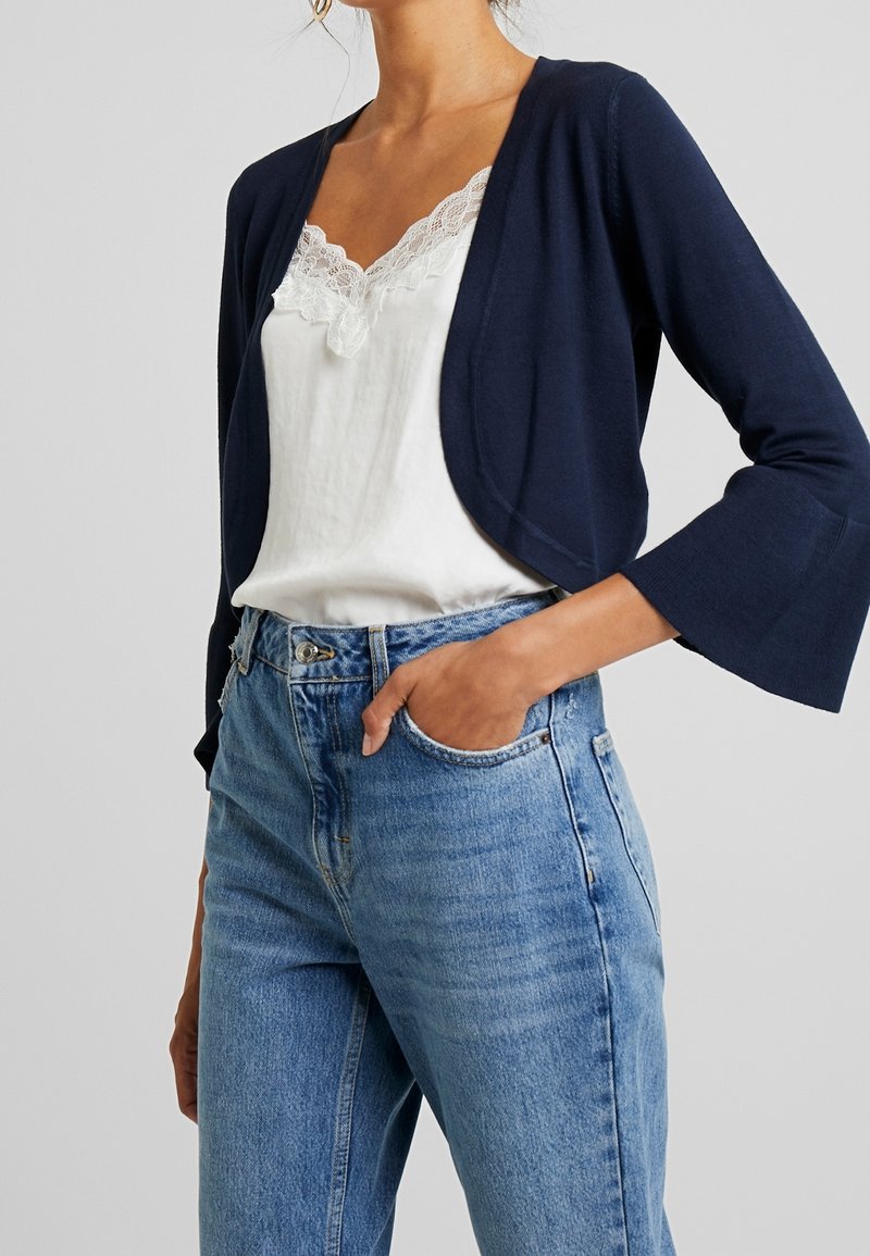 Cardigan court bleu marine avec des manches évasées, associé à un débardeur blanc à bordures en dentelle et un jean en denim bleu clair. Texture lisse, coupe décontractée.