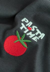 Černá tkanina se vyšitým textem "PASTA TIME" v bílé barvě a červenou rajčatovou ilustrací se zelenými listy. Design má texturovaný povrch.