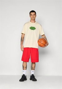 Beige T-skjorte for menn med grønn logo, sammen med lyse røde shorts. Svarte sko og en basketball fullfører det sportslige utseendet.