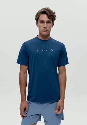 Mladý muž s vlnitými vlasmi v námorníckej modrej športovej košeli s krátkymi rukávmi a svetlomodrých športových šortkách, stojaci pred jednoduchým pozadím.