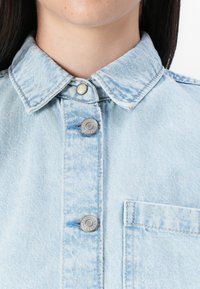 A close-up of a person's neck and collar wearing a light blue denim jacket. Features include button details and a chest pocket.