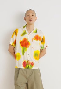 Camicia a maniche corte con bottoni in tessuto crema, caratterizzata da un motivo floreale in tonalità arancione, rosso e verde, con taschino sul petto e colletto.