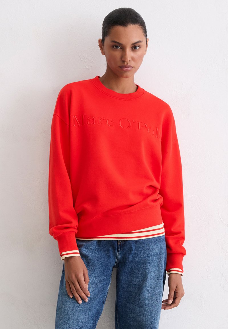 Czerwona bluza z logo "Marc O’Polo" wytłoczonego, z ściągaczami przy mankietach i paskiem w paski. Połączona z niebieskimi dżinsami. Gładka faktura.