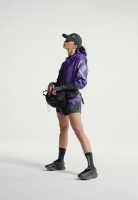 Frau in lila Jacke, schwarzen Shorts, Basecap und Sneakern steht, hält eine schwarze Tasche und blickt nach oben vor einem schlichten Hintergrund.
