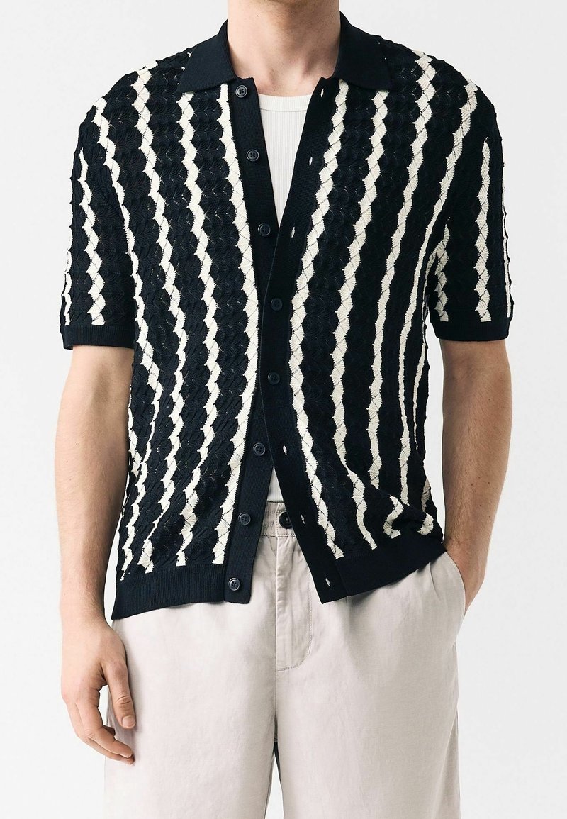 Homme portant une chemise à manches courtes à rayures verticales noires et blanches par-dessus un t-shirt blanc, avec un pantalon beige clair.