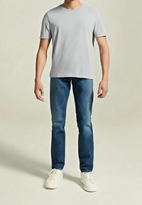 Homme debout portant un t-shirt gris clair uni à manches courtes, un jean bleu et des chaussures blanches à lacets, sur un fond neutre.