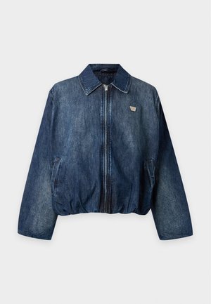 Un homme porte une veste en jean bleue, une chemise blanche, un pantalon beige, des chaussures foncées et une casquette de baseball grise sur un fond uni.
