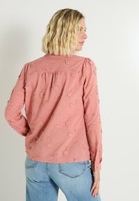 Femme avec des cheveux blonds ondulés portant un blouse à manches longues rose poussiéreux avec des broderies florales et un jean bleu clair, de profil contre un mur blanc.