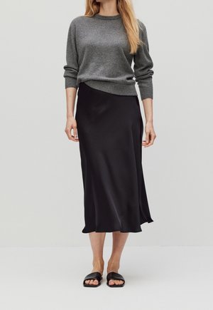 Femme portant un pull gris à manches longues, une jupe midi en soie noire et des sandales slides noires sur un fond clair uni.