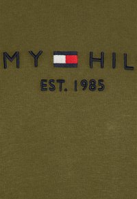 T-shirt in cotone verde oliva con testo ricamato "MY HIL" e un logo rettangolare nei colori rosso, bianco e blu; data di fondazione "EST. 1985."