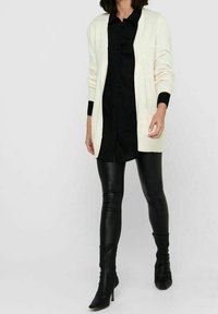 Krämfärgad cardigan med långa ärmar och fickor, bärs över en svart skjorta med knappar och svarta läderleggings, tillsammans med svarta klackstövlar.