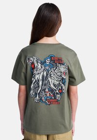 Camiseta de manga corta verde oliva con un gráfico de dos perros feroces, un esqueleto y el texto "Ride the Rails", con detalles intrincados.