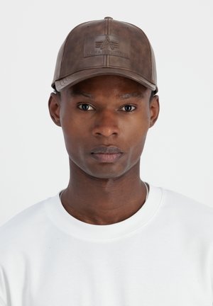 Joven de piel oscura que lleva una gorra de béisbol de cuero marrón con el logo "Alpha Industries" grabado y una camiseta blanca de cuello redondo.