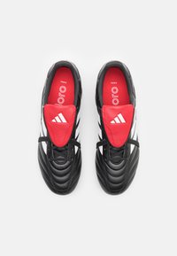Des chaussures de football noires avec un dessus texturé, présentant une languette rouge, des lacets noirs et trois rayures blanches de chaque côté.